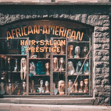 Showcase of various wigs in a store window labeled 'African-American Hair Saloon Prestige'.